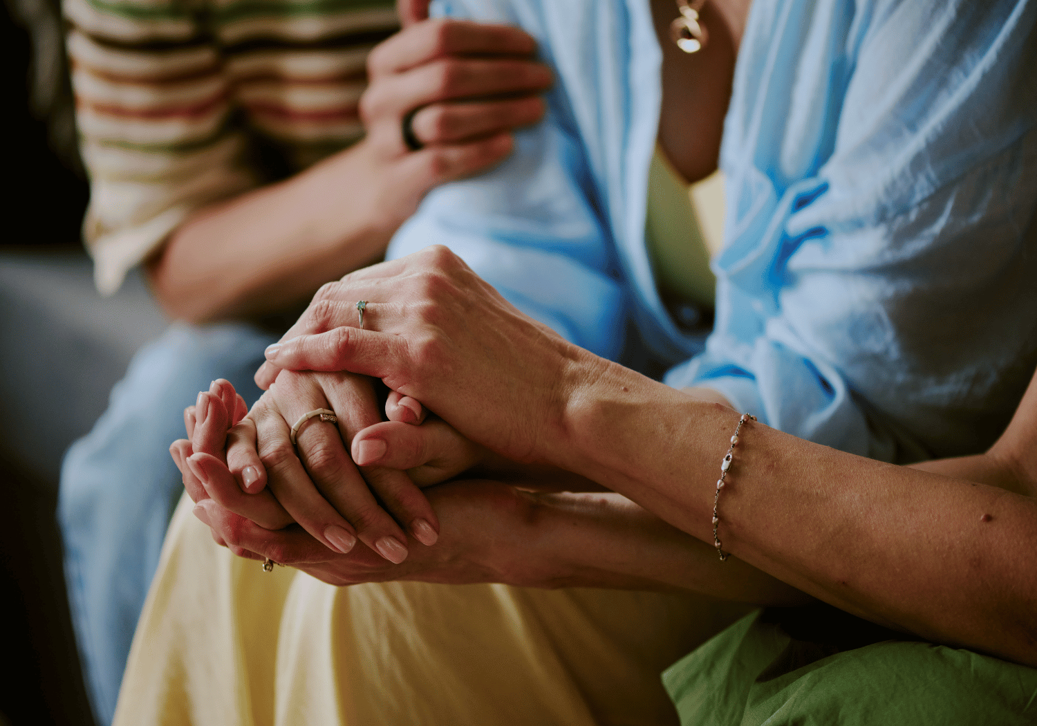 Two people hold hands in a supportive, comforting gesture; multiple rings and a bracelet visible on the wrists and fingers.
